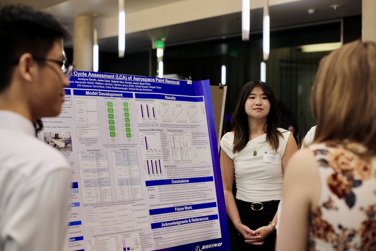Students stand by their poster and listen to a guest at the Capstone Showcase reception. 