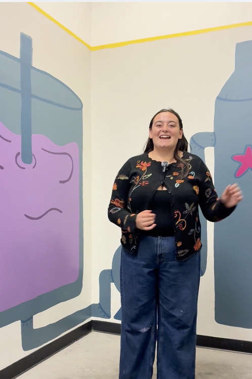 Naudia Watkins stands in front of a mural in Benson that she painted. Behind her, the mural depicts a large container stirring a purple liquid.