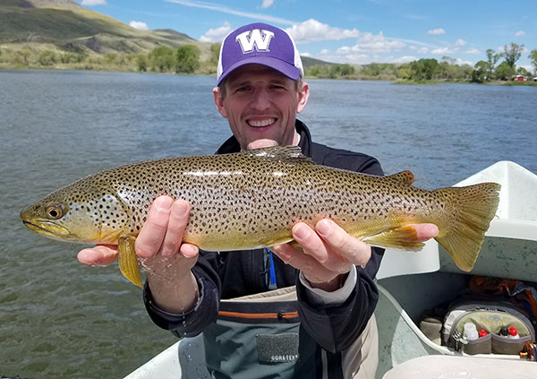 Andy Walker wearing a purple UW hat, standing in front of a lake and holding up a fish