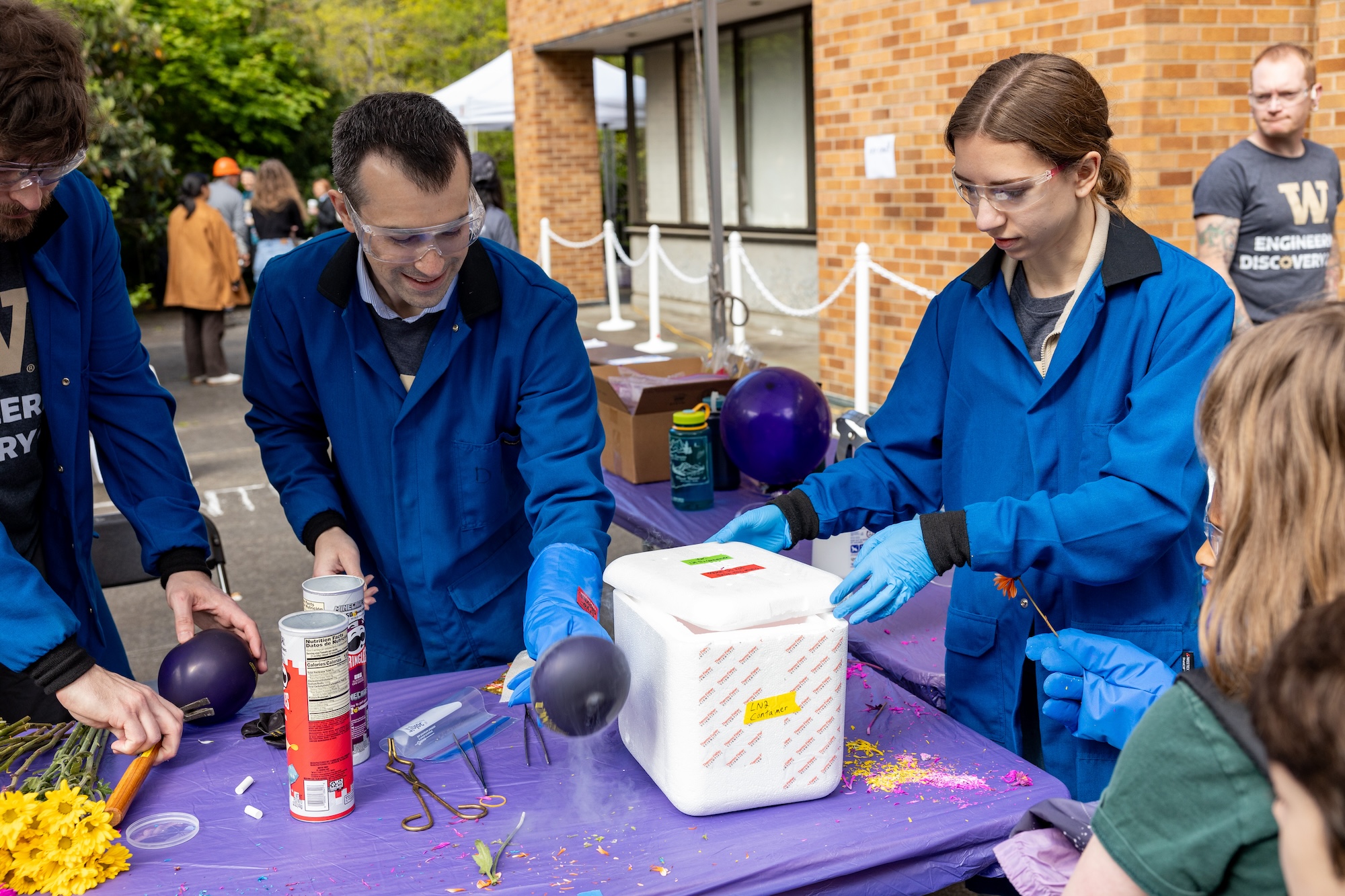 Two people wearing blue lab coats, gloves and safety goggles perform a science demonstration for a crowd at a youth event
