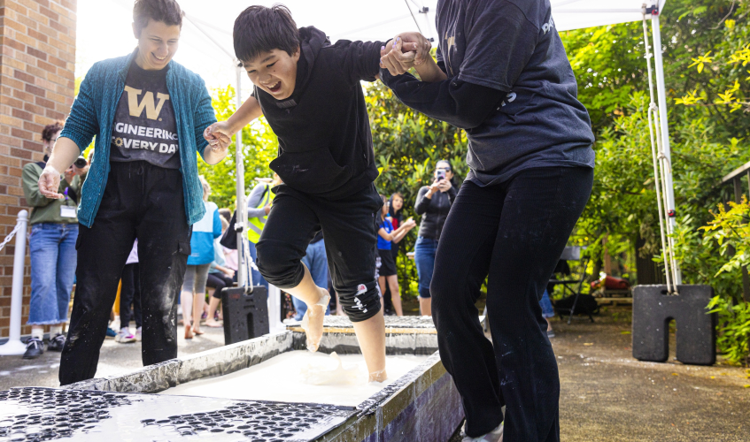 A child laughs while running across a pool of opaque liquid. Two adults stand on either side of the child and hold his hands.