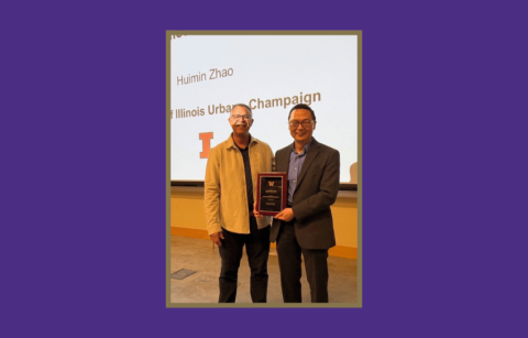 Two people stand in front of a large powerpoint screen. One is holding a wood plaque with gold writing. 