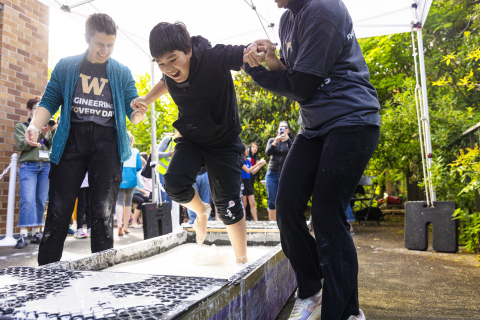 A child laughs while running across a pool of opaque liquid. Two adults stand on either side of the child and hold his hands.