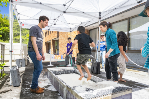 Two ChemE volunteers help a young student jumping in the trough of Oobleck. The student hovers in the air over the pool.