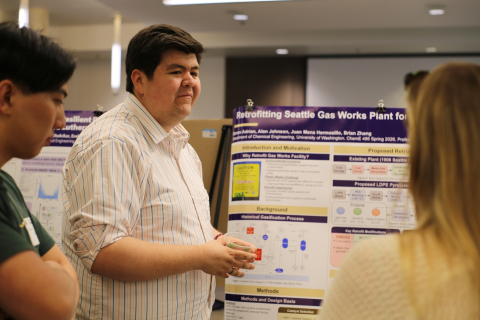 A student stands in front of their research poster, explaining their work to a person standing across from them. 