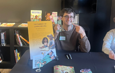 Comes sits at a table at a book release event with copies of his book and a poster explaining the book. 