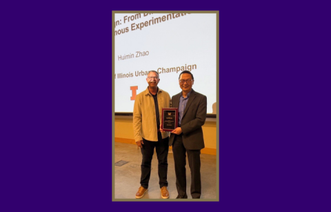 Two people stand in front of a large projector screen. One is holding a plaque with a wood base and gold writing.