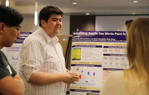 A student stands in front of their research poster explaining their research to someone standing across from them. 